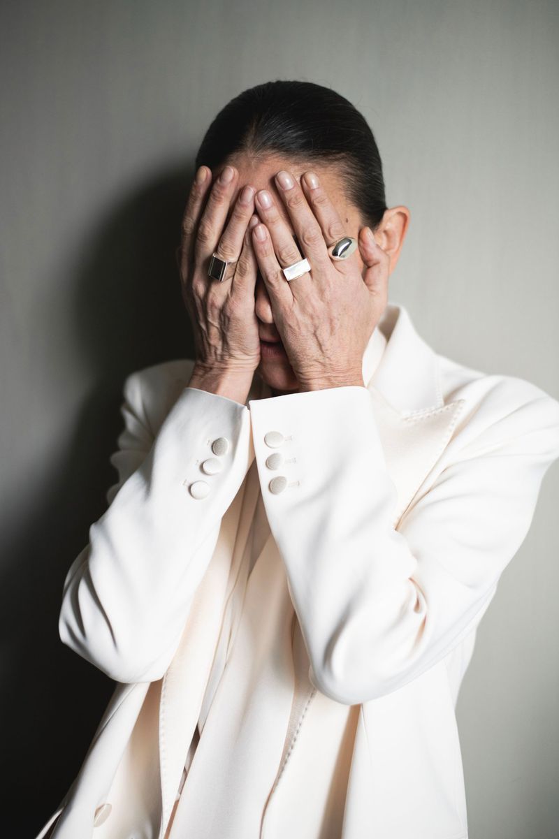 Mature adult covers face with hands wearing a white blazer and several rings against a neutral background in an eye-level medium shot.