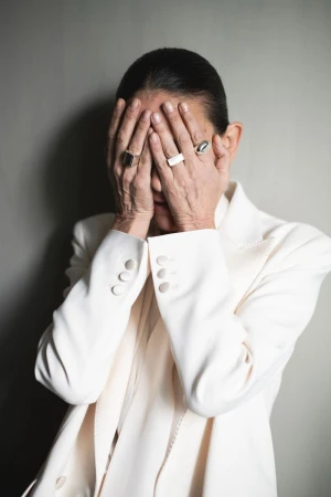 Mature adult covers face with hands wearing a white blazer and several rings against a neutral background in an eye-level medium shot.