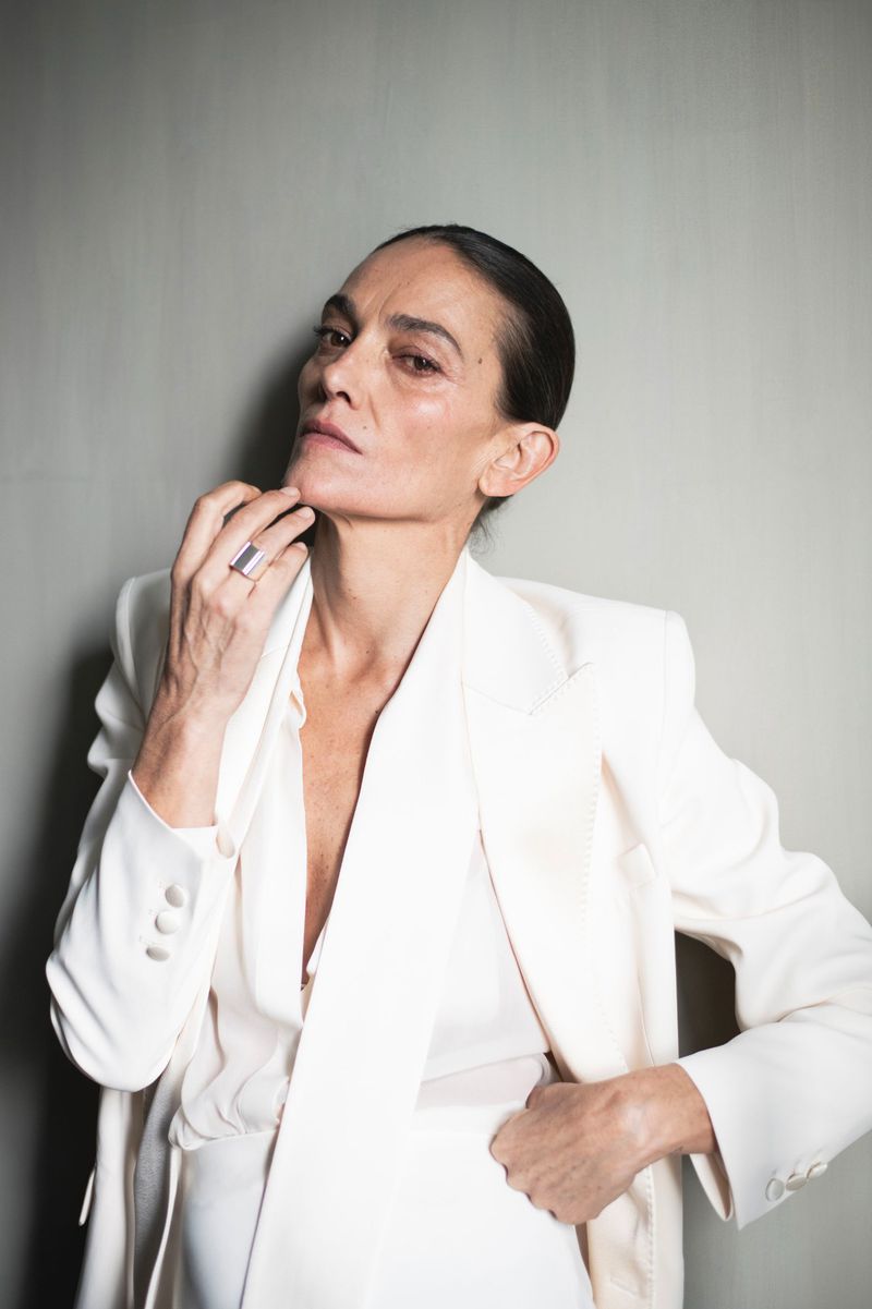 Elegant adult woman poses in a white blazer and top in a studio setting with gray background, showcasing fashion and beauty.