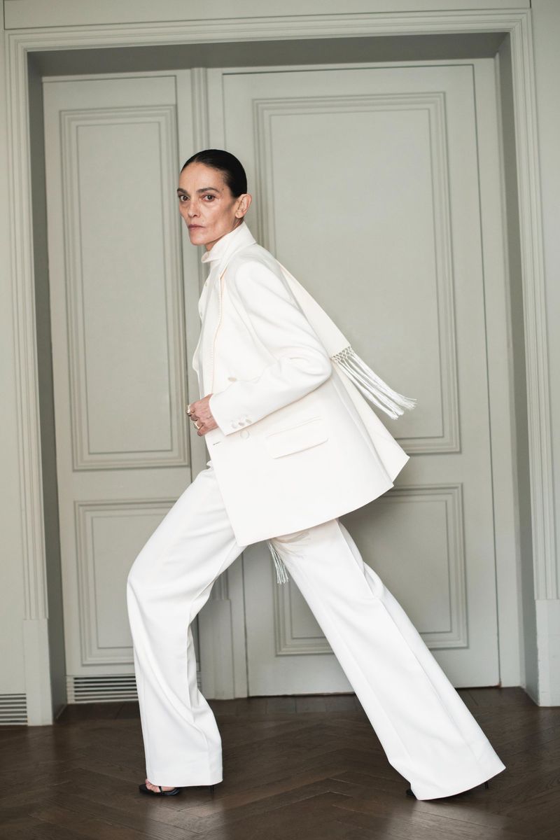 Woman in all-white suit with wide-leg pants, posing in front of grey paneled doors, fashion editorial style, neutral indoor lighting.