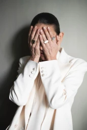 Model Laura Ponte covering face with hands wearing white blazer and silver rings against grey background
