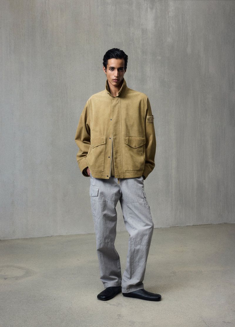 A young adult male poses in a full shot, wearing a tan jacket and grey cargo pants against a neutral studio backdrop.