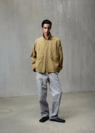 A young adult male poses in a full shot, wearing a tan jacket and grey cargo pants against a neutral studio backdrop.