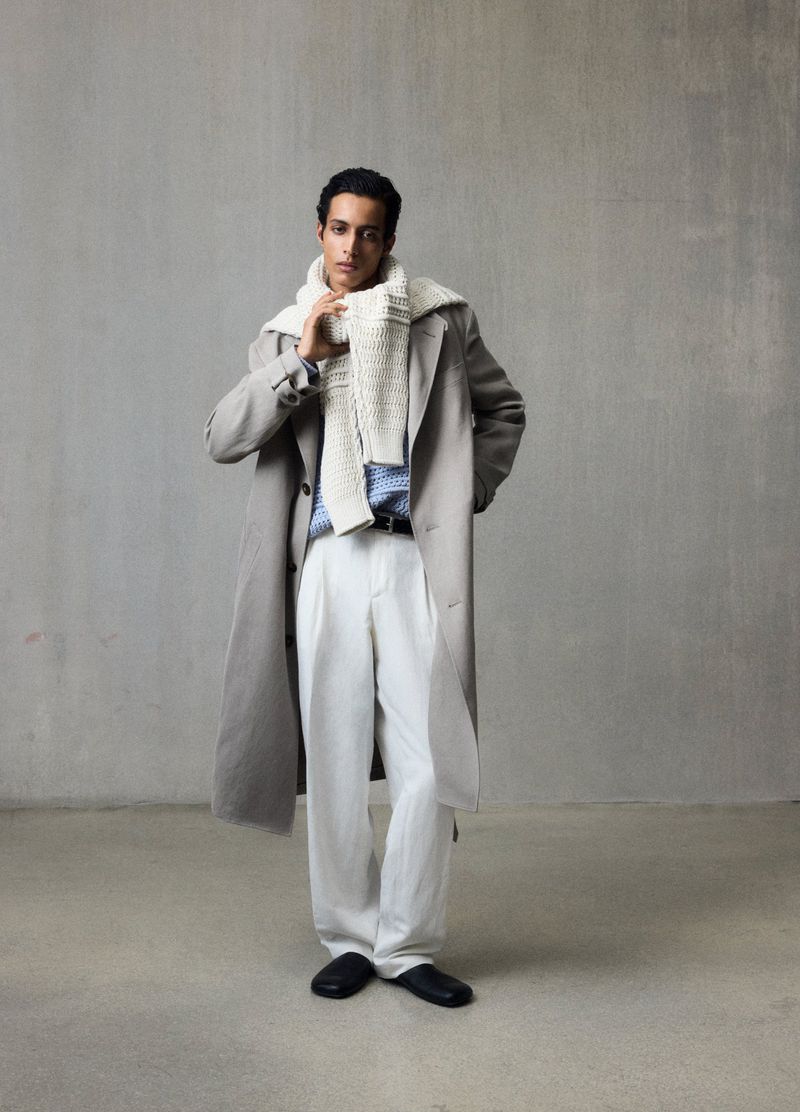A stylish dark-haired man models a light-grey trench coat, knit scarf, white pleated pants, and black shoes in a full-body studio shot.