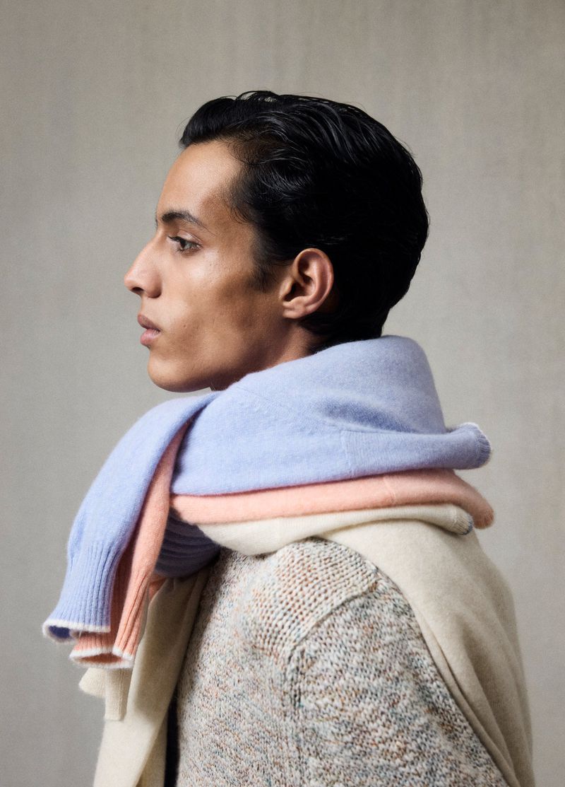 Profile view of a man draped with pastel cashmere sweaters. He wears a tweed coat against a neutral background in a fashion-focused shot.