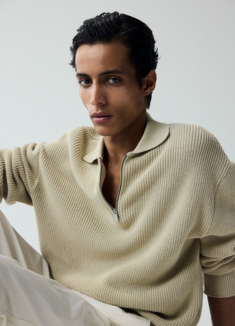 Close-up of a young adult male posing in a studio wearing a beige knitted sweater with a zipper and light-colored pants.