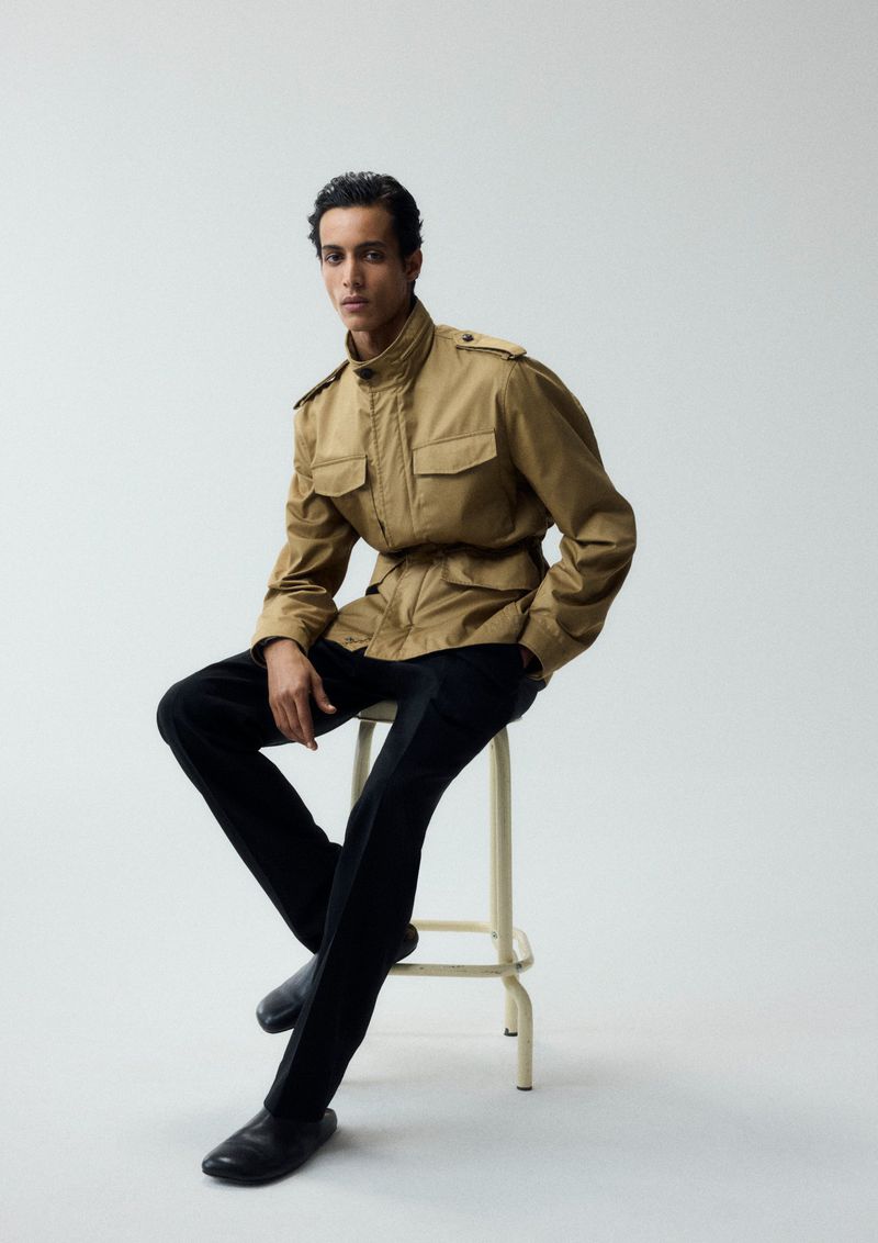 Man wearing a tan field jacket and black trousers sitting on a simple metal stool against a plain light grey background.