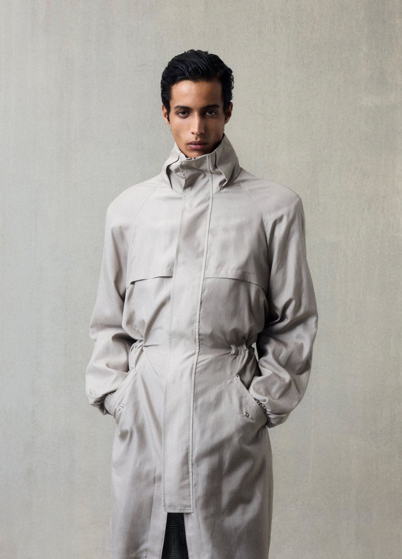 Indoor portrait of a young adult man wearing a long, light-gray trench coat looking directly at the camera against a neutral gray backdrop.