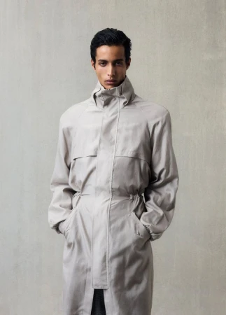 Indoor portrait of a young adult man wearing a long, light-gray trench coat looking directly at the camera against a neutral gray backdrop.