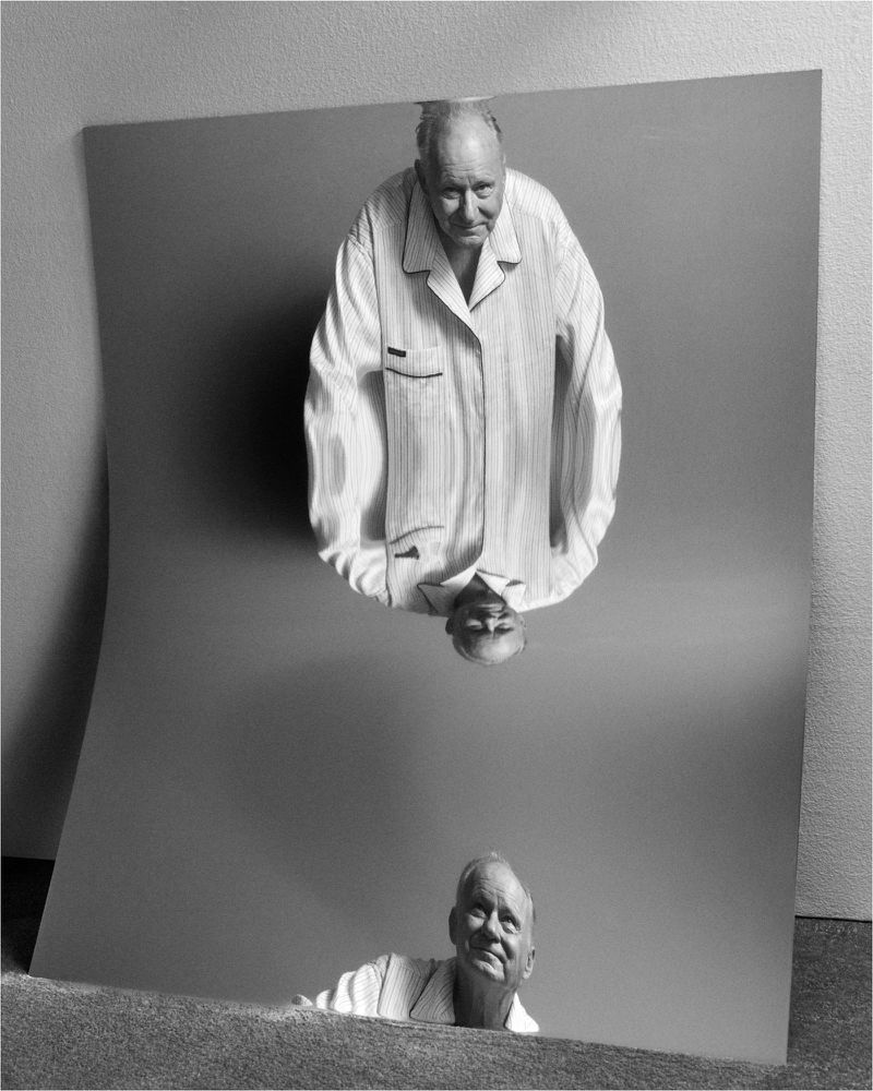 Older man in striped pajamas gazes at his reflection in a slightly warped standing mirror with a gray background in a black and white portrait.