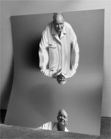 Older man in striped pajamas gazes at his reflection in a slightly warped standing mirror with a gray background in a black and white portrait.