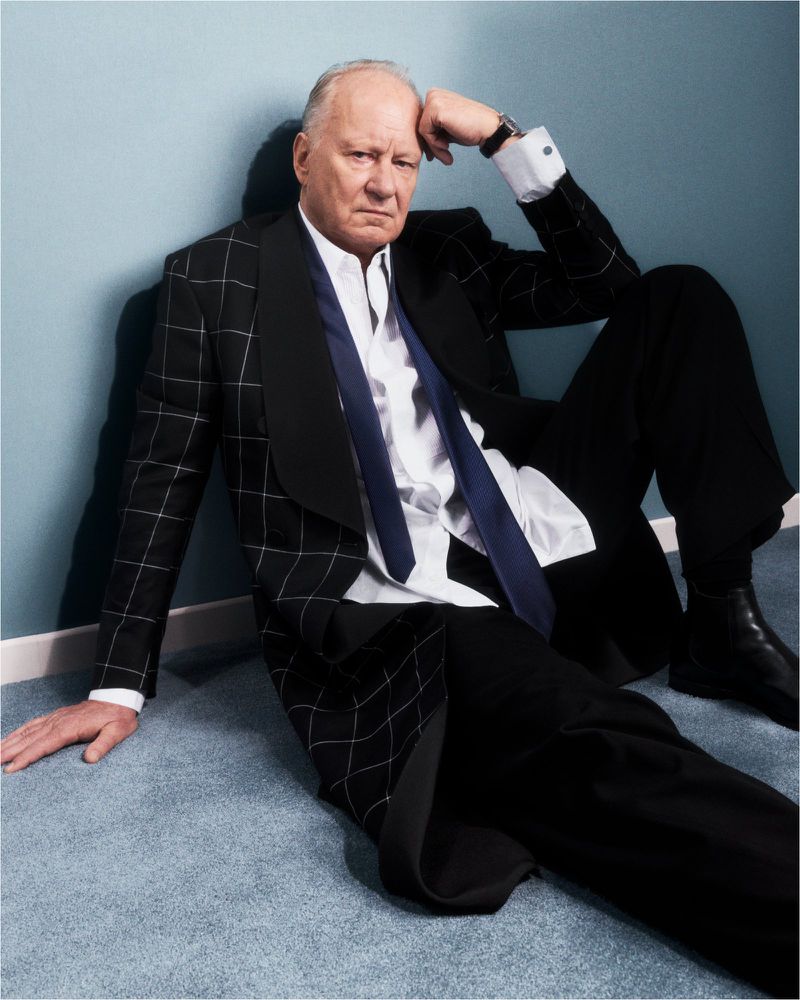 Older man in black suit and tie sits against a blue wall. He rests his head on his hand in a thoughtful pose indoors.