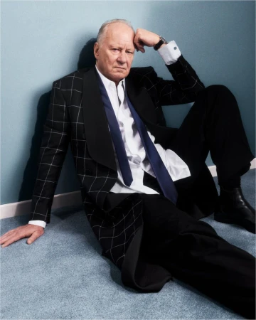 Older man in black suit and tie sits against a blue wall. He rests his head on his hand in a thoughtful pose indoors.