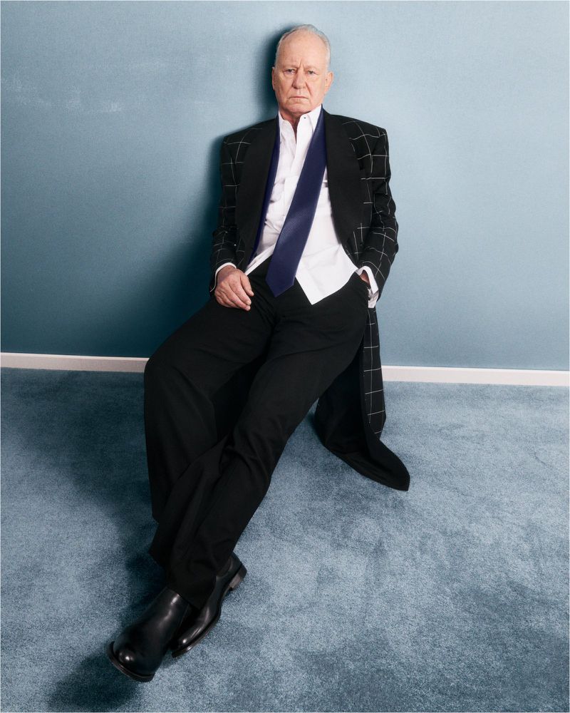 Older adult man in a dark suit and blue tie, seated against a blue wall with a carpeted floor, looking directly at the camera.