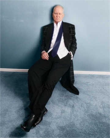 Older adult man in a dark suit and blue tie, seated against a blue wall with a carpeted floor, looking directly at the camera.