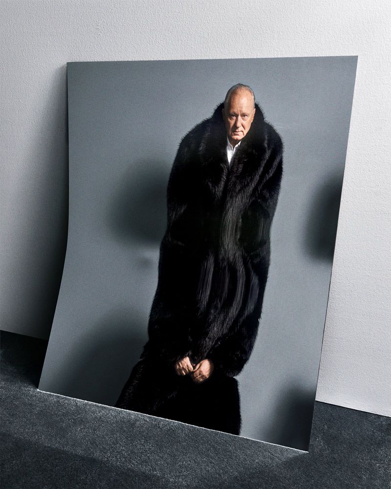 An older adult male in a black fur coat poses against a grey backdrop, presented as a photograph leaning against a dark surface.