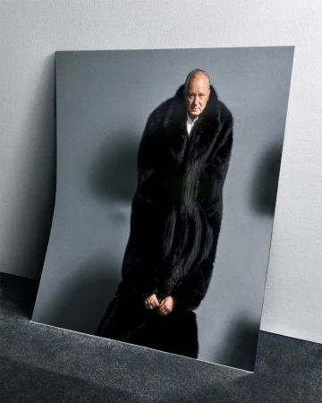 An older adult male in a black fur coat poses against a grey backdrop, presented as a photograph leaning against a dark surface.