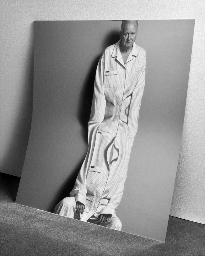 Black and white studio portrait of an adult posing on a sheet of folded paper, wearing striped pajamas against a neutral background.