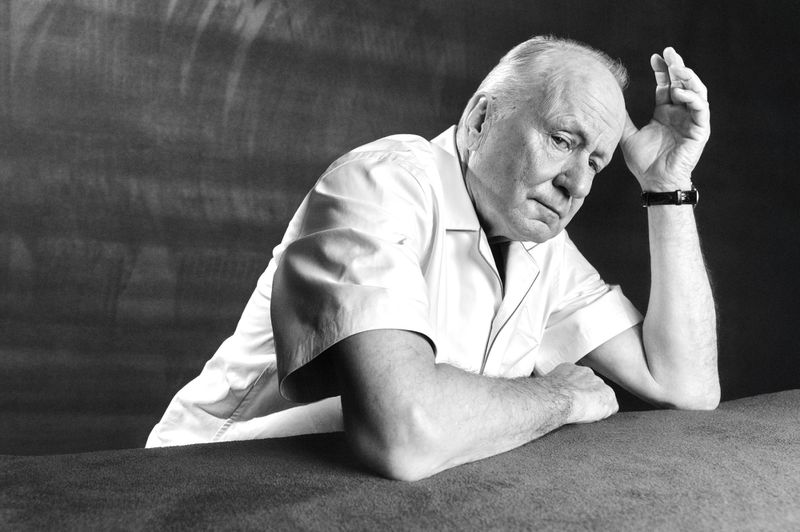 Black and white portrait of an older man with white hair wearing a short-sleeved collared shirt resting his head on his hand.