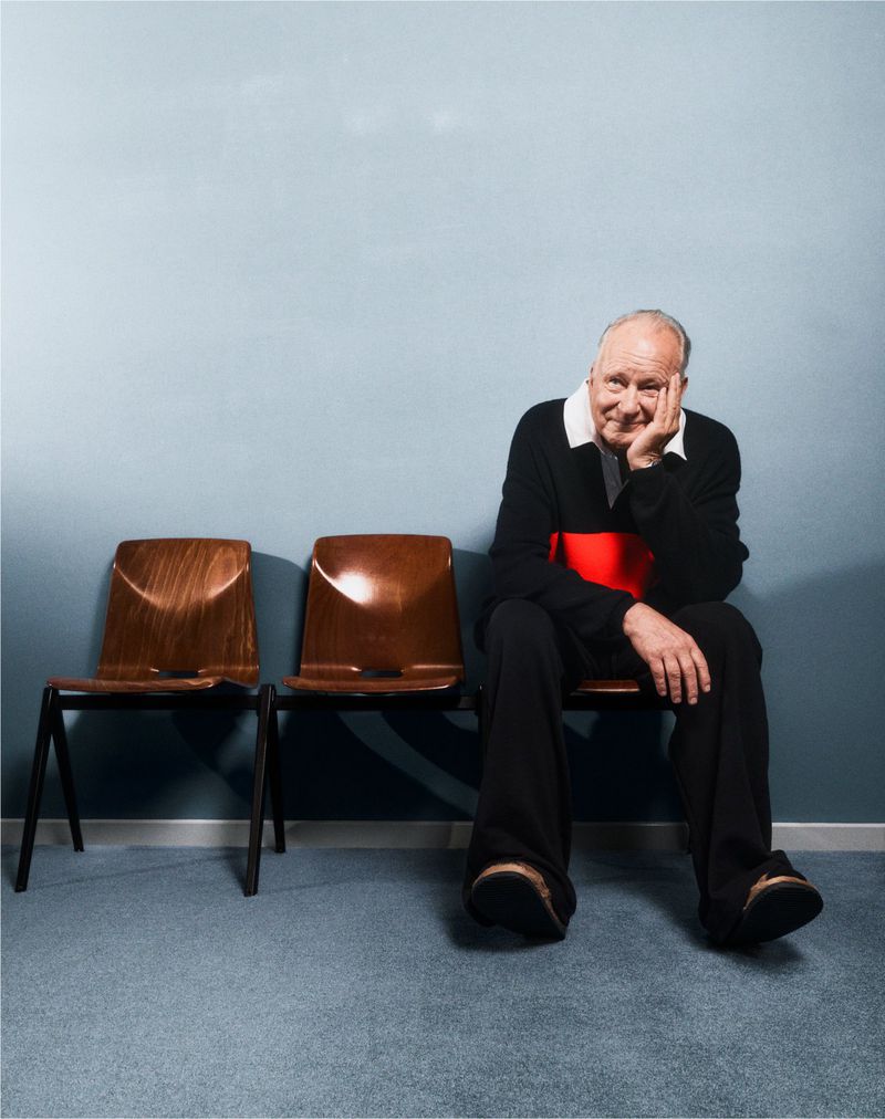 Elderly man sits on a brown chair in an indoor setting. The man smiles with his hand to his face, wears a black sweater.
