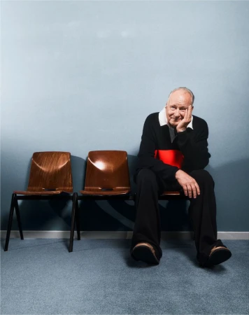 Elderly man sits on a brown chair in an indoor setting. The man smiles with his hand to his face, wears a black sweater.