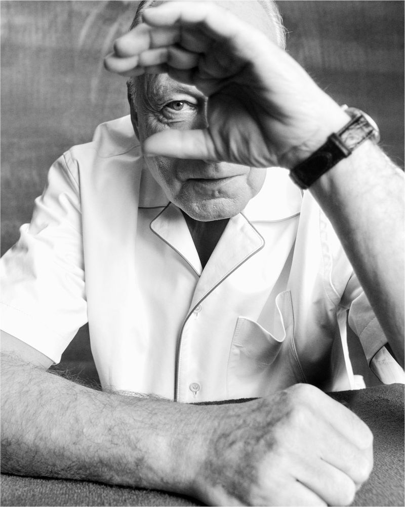 Portrait of an elderly man shielding his face with his hand, wearing a white collared shirt and a watch, in a black and white photo.
