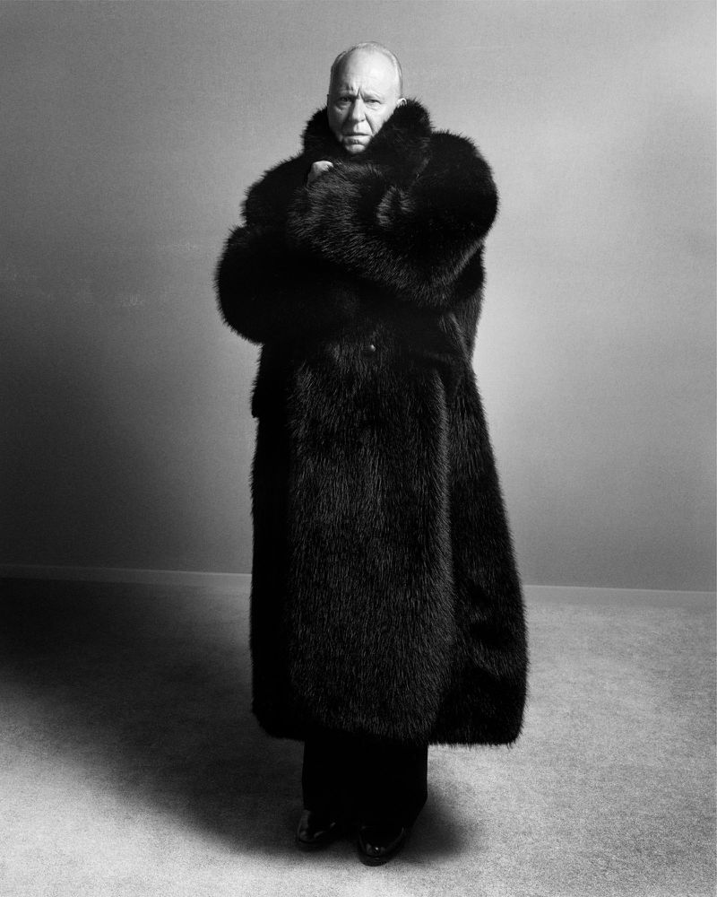 A full-length black-and-white portrait features a bald adult male with arms crossed, wearing a black fur coat against a plain gray background.