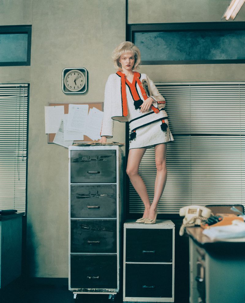 Fashion photo of a blonde model in a white mini dress and jacket, standing on a filing cabinet in an office setting with neutral tones.
