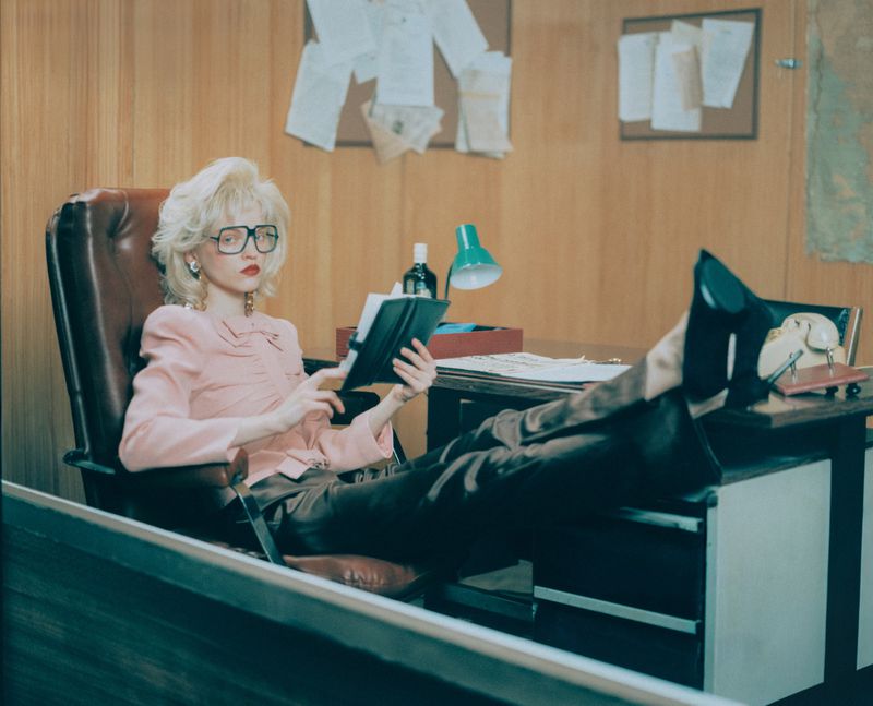 Young woman in pink blouse and brown slacks reclines with feet up on a desk in an office reading a journal, papers pinned to the wall.