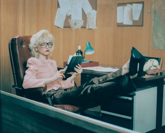 Young woman in pink blouse and brown slacks reclines with feet up on a desk in an office reading a journal, papers pinned to the wall.