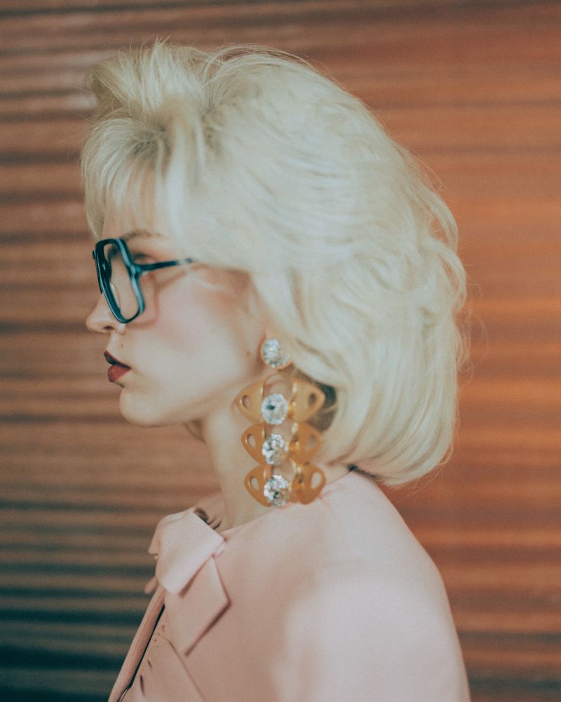 Profile shot of a woman with blond hair, glasses, statement earrings, and a pink blazer, set against a blurred orange background.