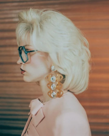 Profile shot of a woman with blond hair, glasses, statement earrings, and a pink blazer, set against a blurred orange background.