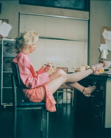 Stylish woman in pink blazer seated at a desk with her feet up, writing, in an office setting with vintage telephone and blinds.