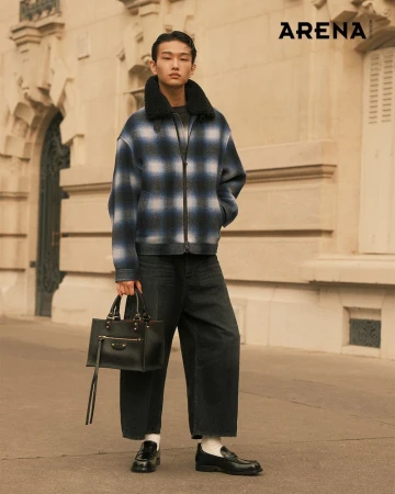 A young adult male in a plaid jacket and wide-leg jeans stands on a city street holding a black leather bag, with an 'ARENA' logo visible.