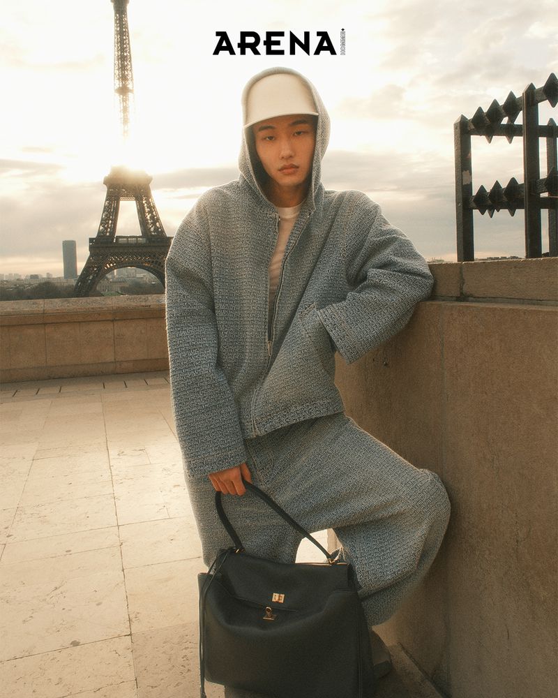 Man in hooded jacket and pants standing in Paris, France, holding a black bag, with the Eiffel Tower in the background.