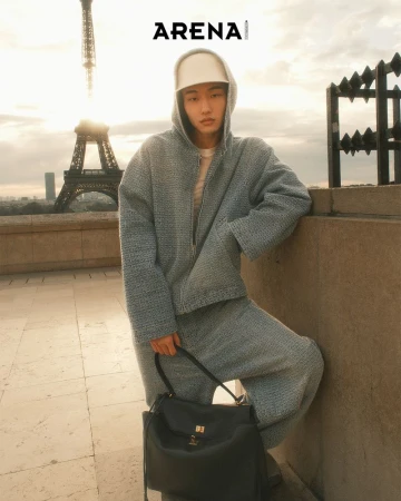 Man in hooded jacket and pants standing in Paris, France, holding a black bag, with the Eiffel Tower in the background.