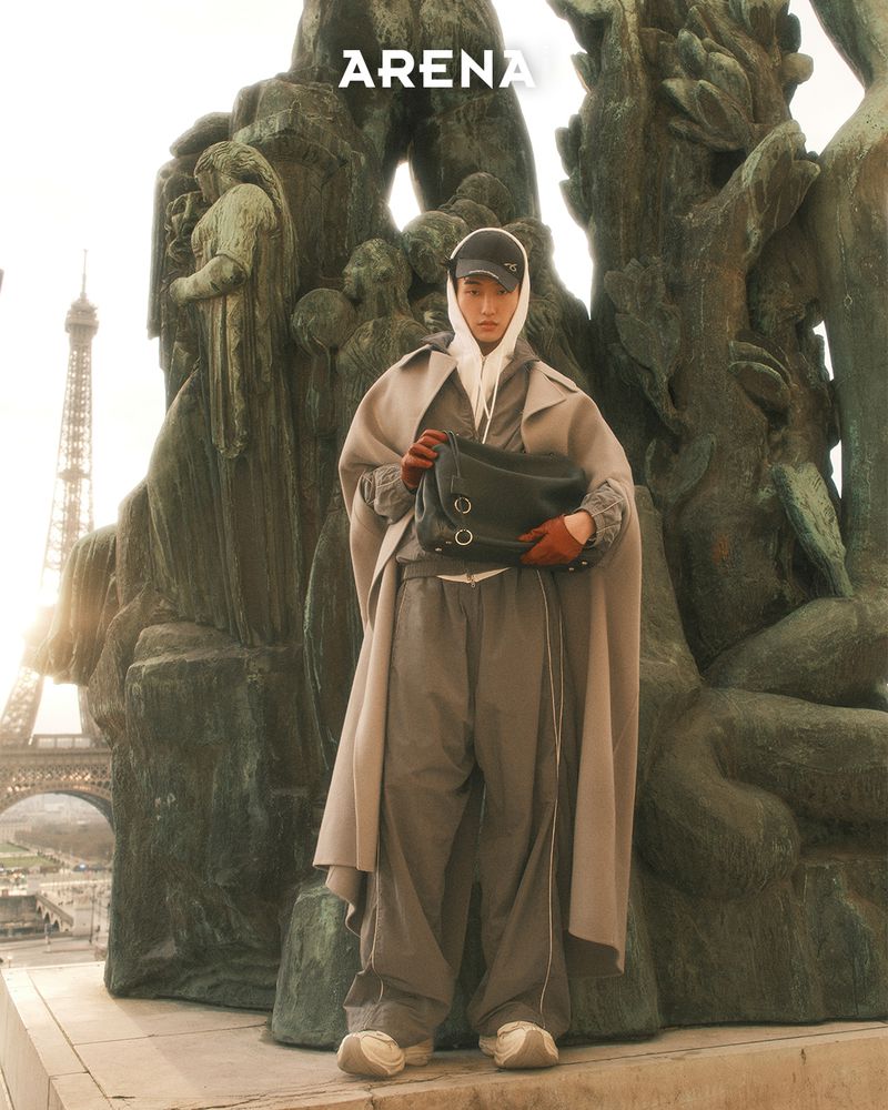 Fashion shot featuring a male model in streetwear-inspired clothing and a shoulder bag, posing in front of a Parisian statue and the Eiffel Tower.
