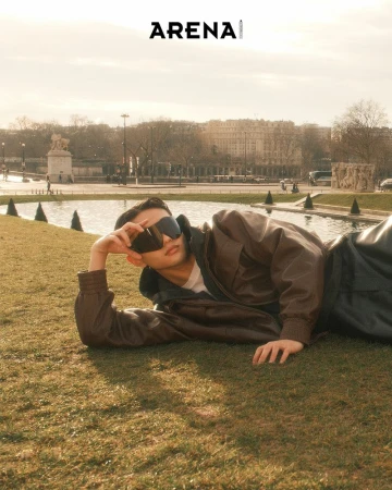 Adult male model in sunglasses poses on grass in an outdoor editorial fashion layout with city buildings and sky in the background.