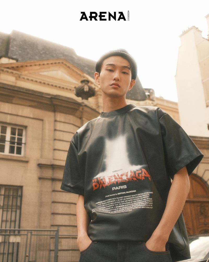 Adult male in an outdoor urban environment, wearing a dark graphic t-shirt, jeans and beanie, stands against a light stone building.