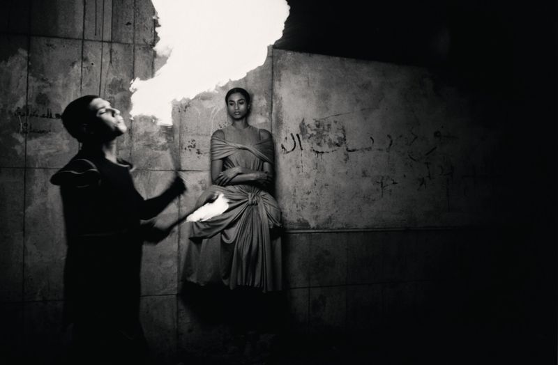 Monochrome photograph featuring an androgynous adult blowing fire as a woman in draped fabric sits against a textured wall.