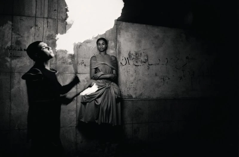 Monochrome photograph featuring an androgynous adult blowing fire as a woman in draped fabric sits against a textured wall.