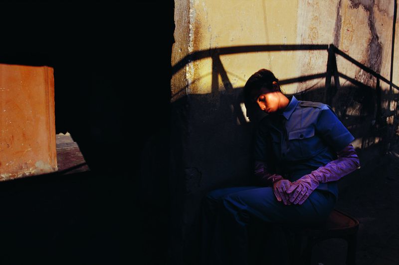 Imaan Hammam seated against a textured wall wearing blue uniform shirt and trousers with dramatic shadows.