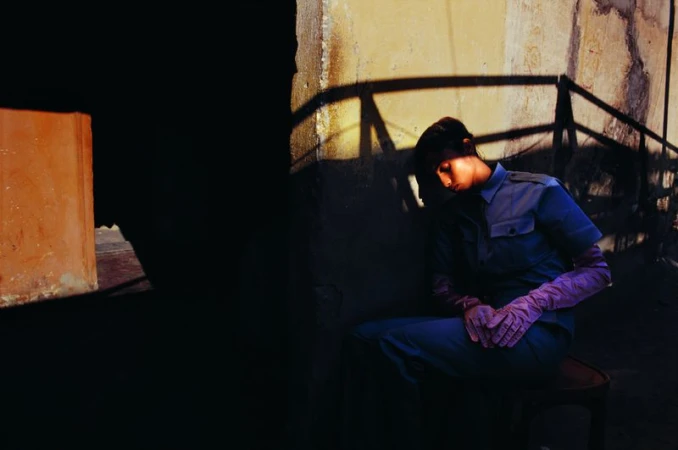 Imaan Hammam seated against a textured wall wearing blue uniform shirt and trousers with dramatic shadows.