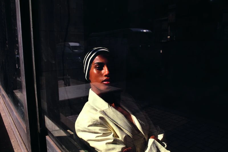 Indoor portrait of a dark-skinned adult woman wearing a striped cap and a light yellow blazer near a glass window with a dark background.
