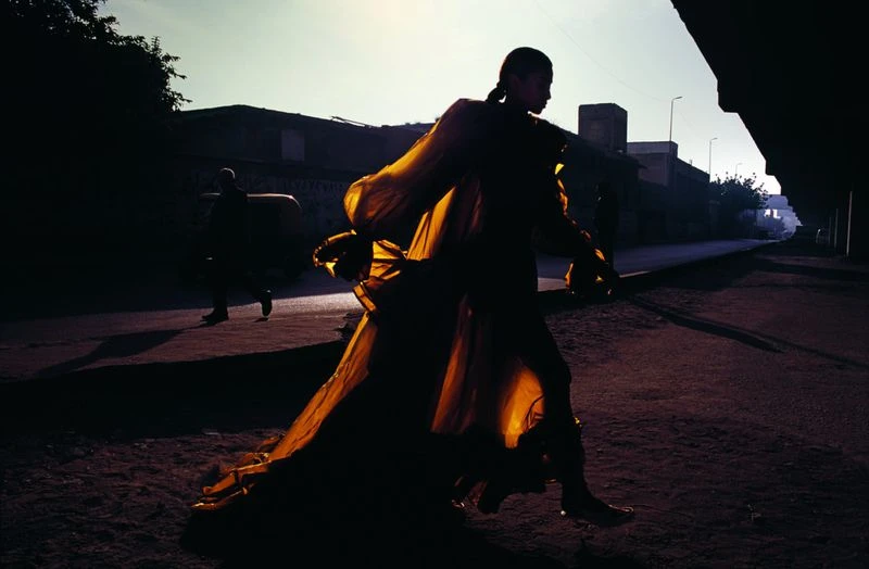 Woman in golden gown walks down deserted street at dusk. Dramatic fashion shot with silhouette and shadows, urban setting with buildings.