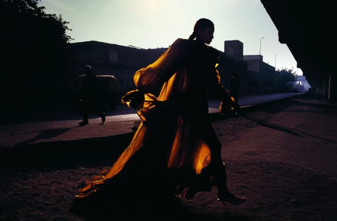 Woman in golden gown walks down deserted street at dusk. Dramatic fashion shot with silhouette and shadows, urban setting with buildings.