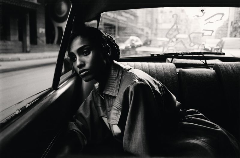 Monochrome portrait of a woman with braided hair, wearing a collared jacket in the backseat of a car, looking out the window at a cityscape.