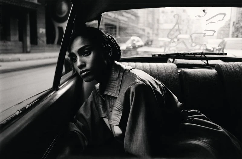 Monochrome portrait of a woman with braided hair, wearing a collared jacket in the backseat of a car, looking out the window at a cityscape.