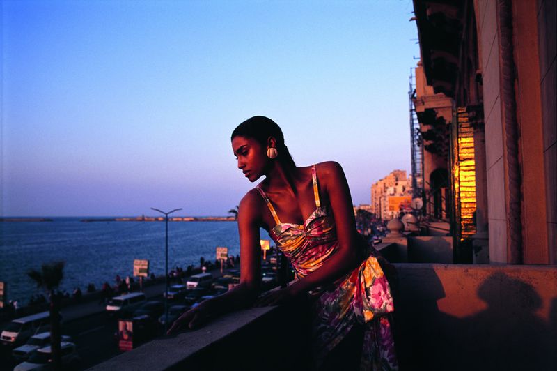 Woman poses on a balcony at dusk wearing a floral dress. Ocean and buildings are visible in the background as the sun sets.