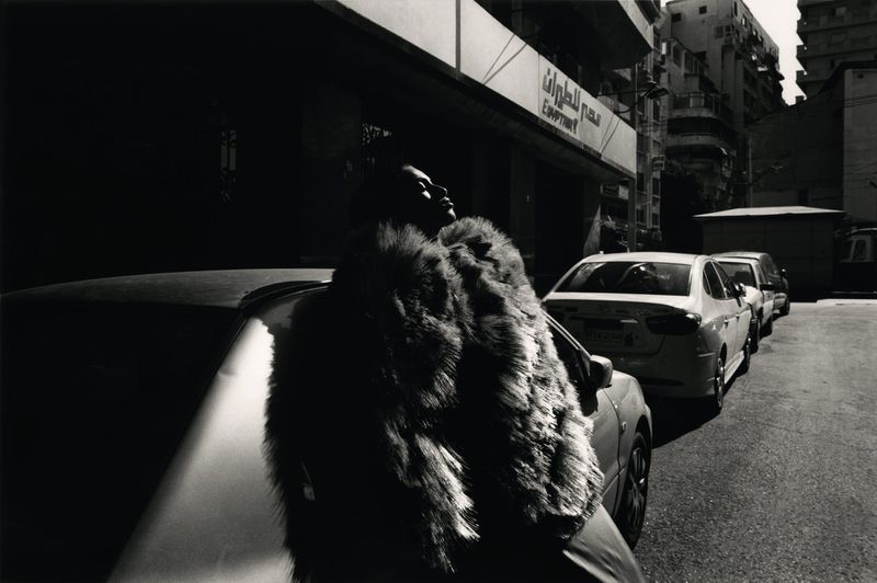 Black and white outdoor shot of an adult wearing a fur coat standing next to a parked car on a city street, with nearby buildings.